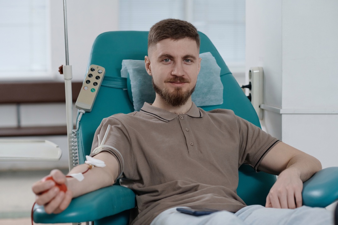 Young caucasian man sitting in armchair in clinic during blood transfusion and smiling at camera